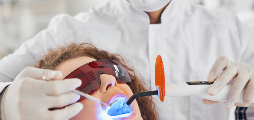 Attractive lady with curly hair lying in dental chair in protective glasses with opened mouth while while male doctor in white glasses doing whitening procedure. Concept of dental care
